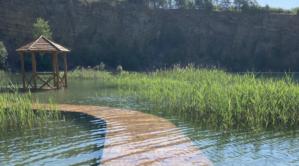 Foto van een vlonder die net onder het wateroppervlak ligt. De vlonder leidt naar een prieeltje midden een prachtig meer. 
