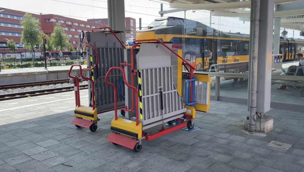 Een foto van twee zogenaamde 'vlinderbruggen' op het perron van een station. Vlinderbruggen zijn uitklapbare oprijplaten, die worden gebruikt om de afstand tussen perron en trein te overbruggen voor reizigers in bijvoorbeeld een rolstoel. Je ziet een trein op de achtergrond. 